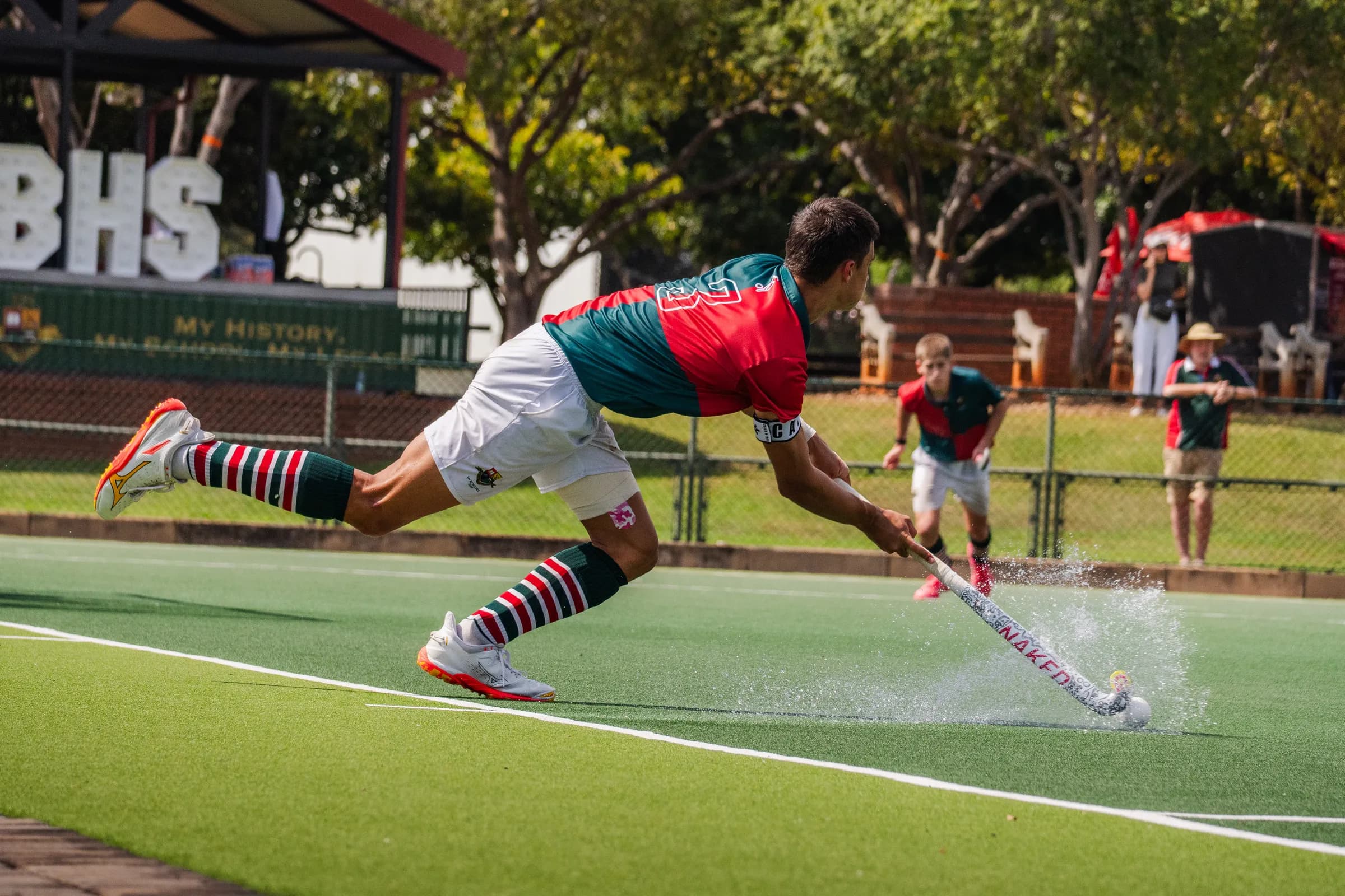 A hockey player lunging full-stretch to strike the ball on the baseline, turf spray flying.