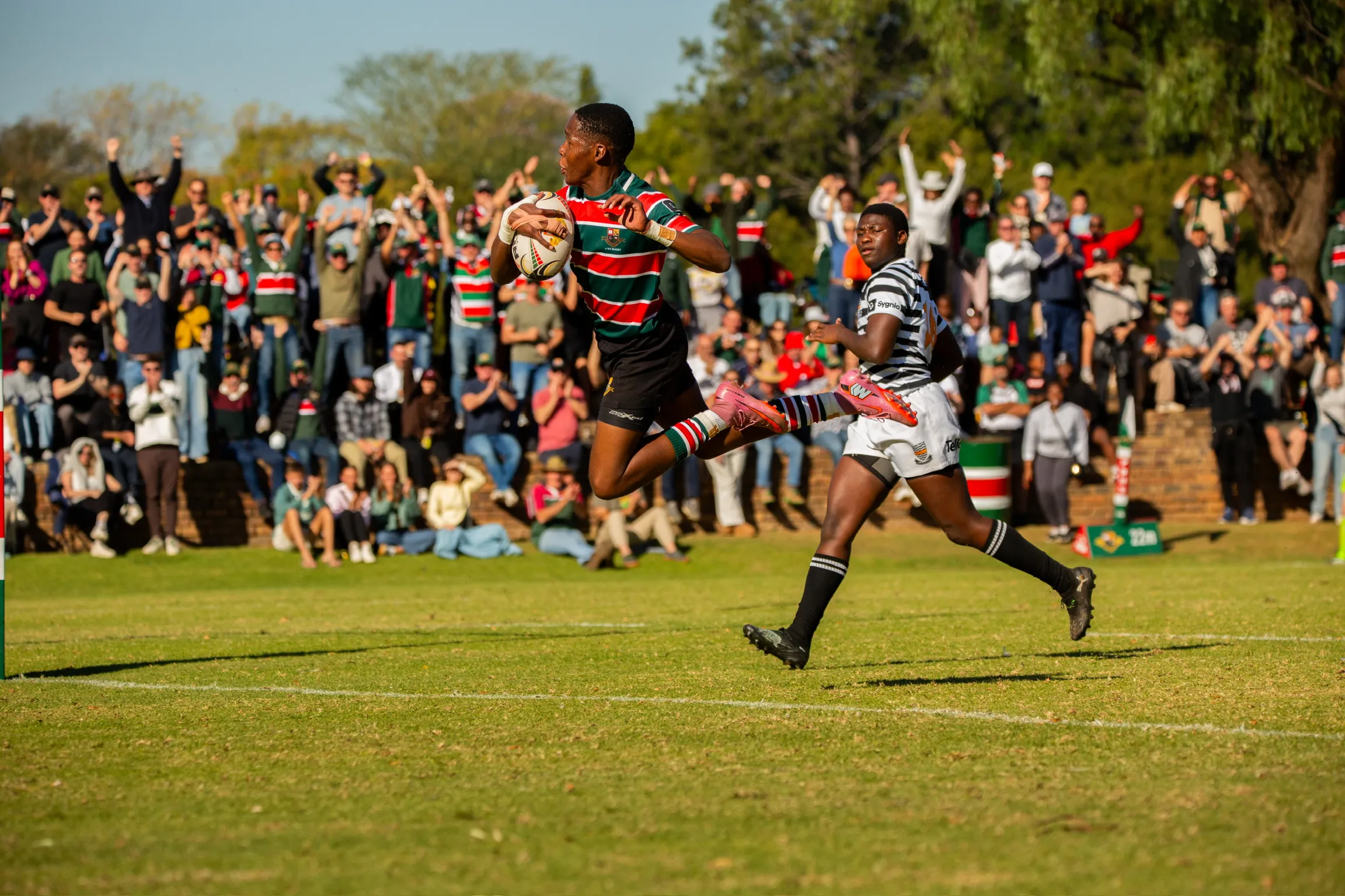 A schoolboy rugby player breaking clear with the ball as the crowd rises behind him.