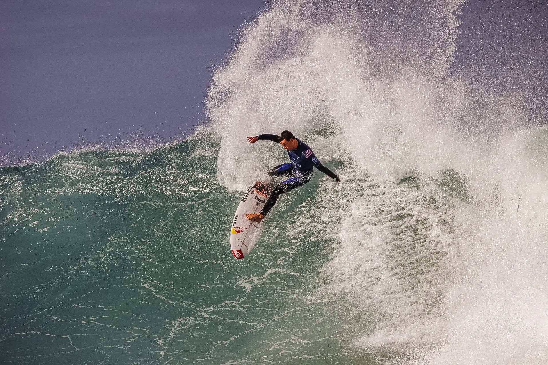 A surfer dropping down the face of a large breaking wave, spray feathering off the lip.