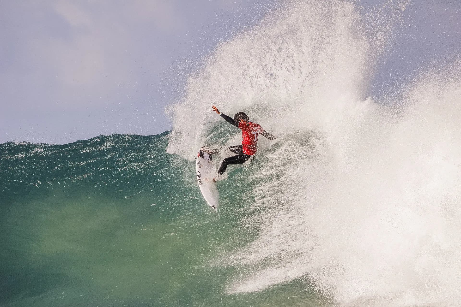 A surfer in a red wetsuit carving hard off the top of a green wall of water.